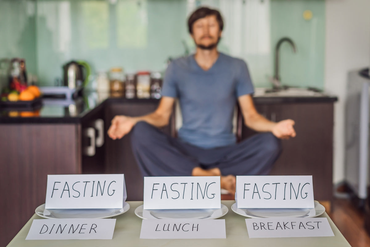 Mature man meditating in his kitchen while three empty plates stand in the forefront with paper place cards saying FASTING, Diner, Lunch and Breakfast.