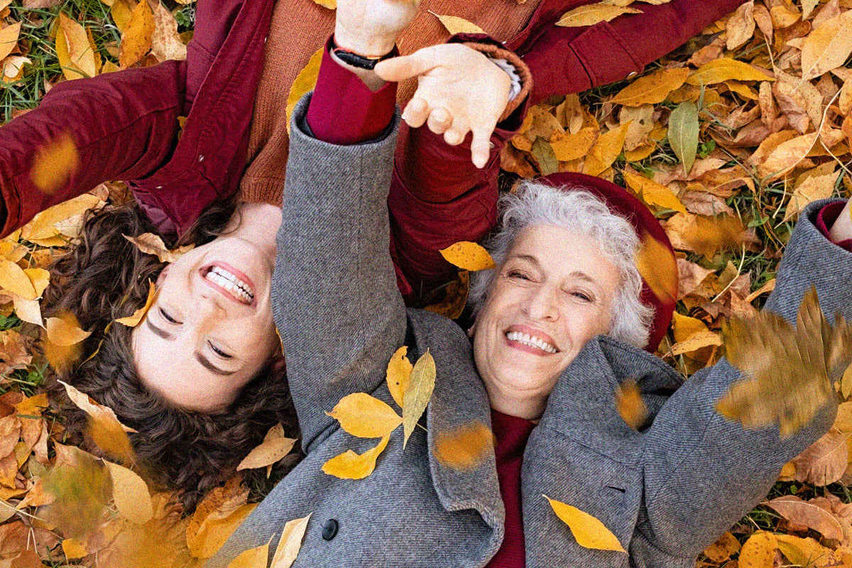 A mature woman and her adult daughter lie laughing with their hands outstretched on a bed of Autumn leaves.