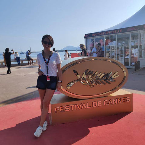 Une femme se tient près du panneau du festival de Cannes avec une belle mer en arrière-plan par une journée ensoleillée.