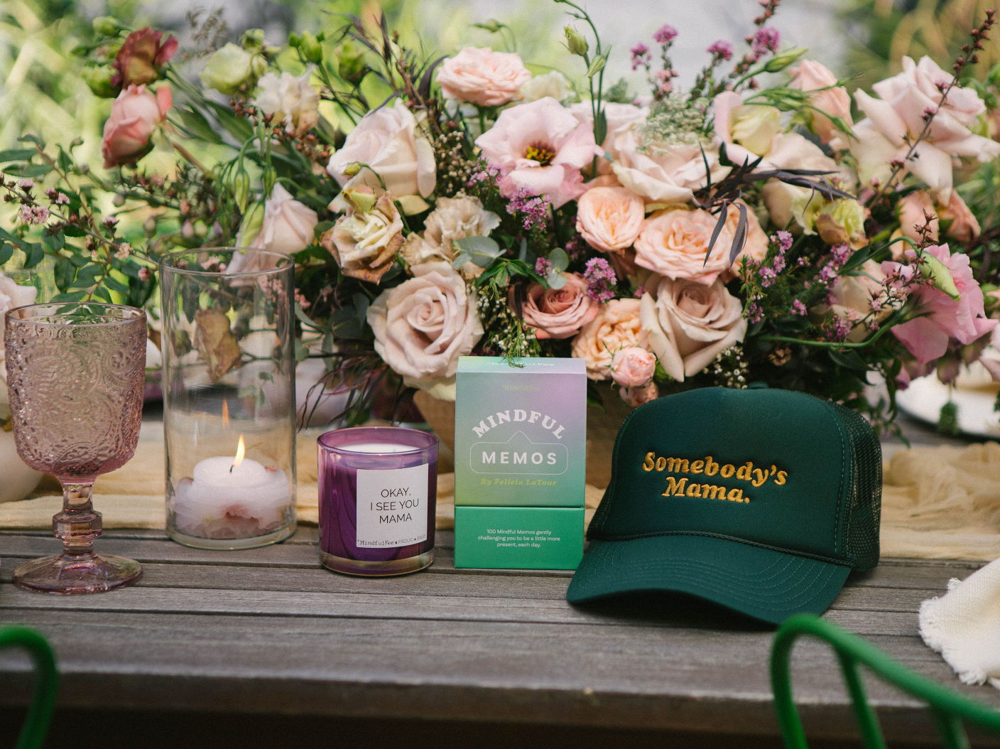 Table decorated with a soft pink flower arrangement, a candle that says, "Okay, I see you Mama", Mindful Memos and a dark green MindfulFee trucker hat