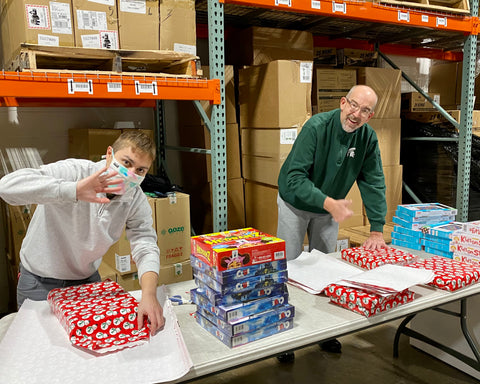 A younger guy and older bald man are standing at a folding table in the Ooze Wholesale warehouse wrapping gifts. They have a pile of games on the table and rolls of wrapping paper, they are actively wrapping the gifts.