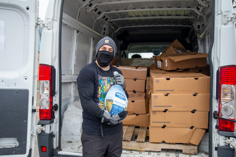 An Ooze Foundation team member wearing black clothes and a black face mask holds up a turkey in front of the trunk of a white van filled with boxes of turkeys.