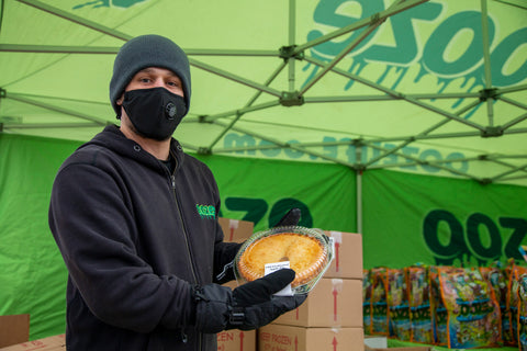 An Ooze Foundation team member in all black, a black hat and black face mask holds up a pie. He is standing under the green Ooze tent.