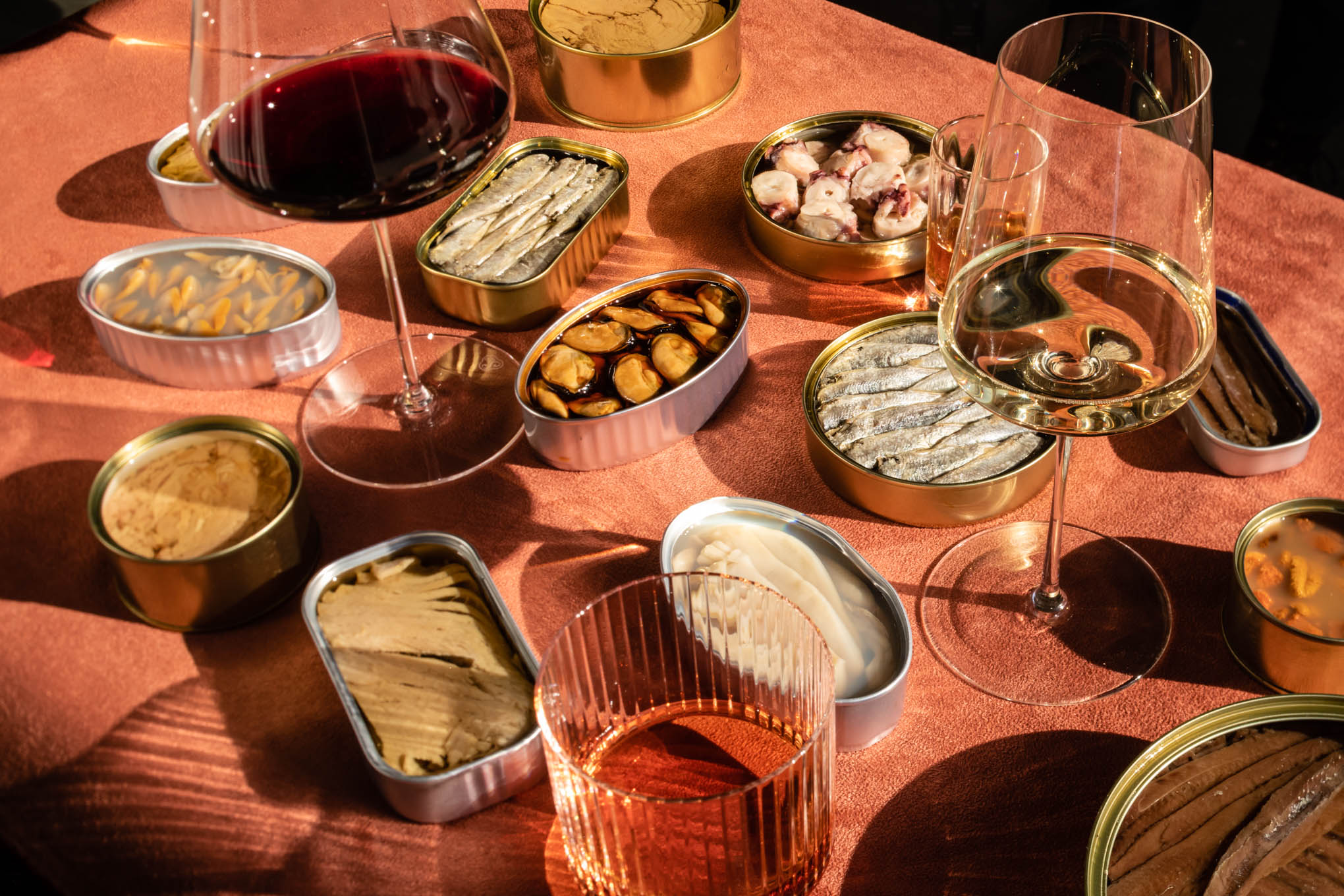 A large variety of seafood conserva cans opened amongst wine glasses on a pink surface