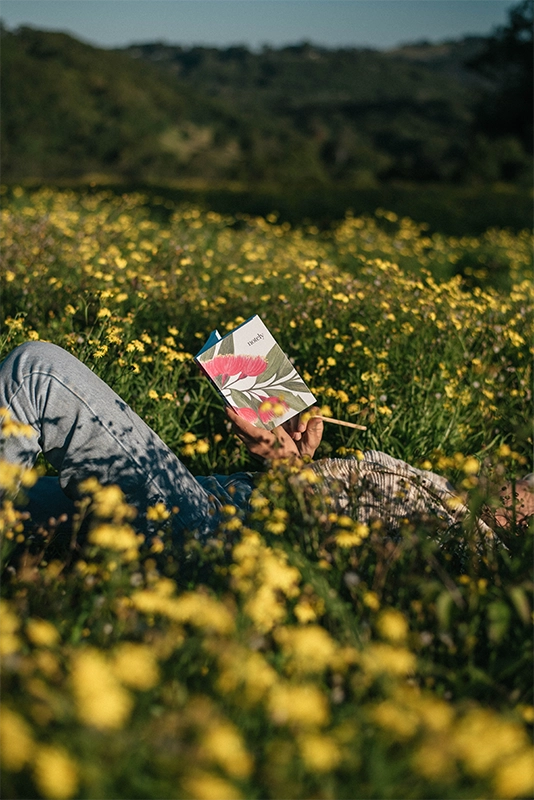 Woman laying in a flower field with the Notely Blossom journal in her hands