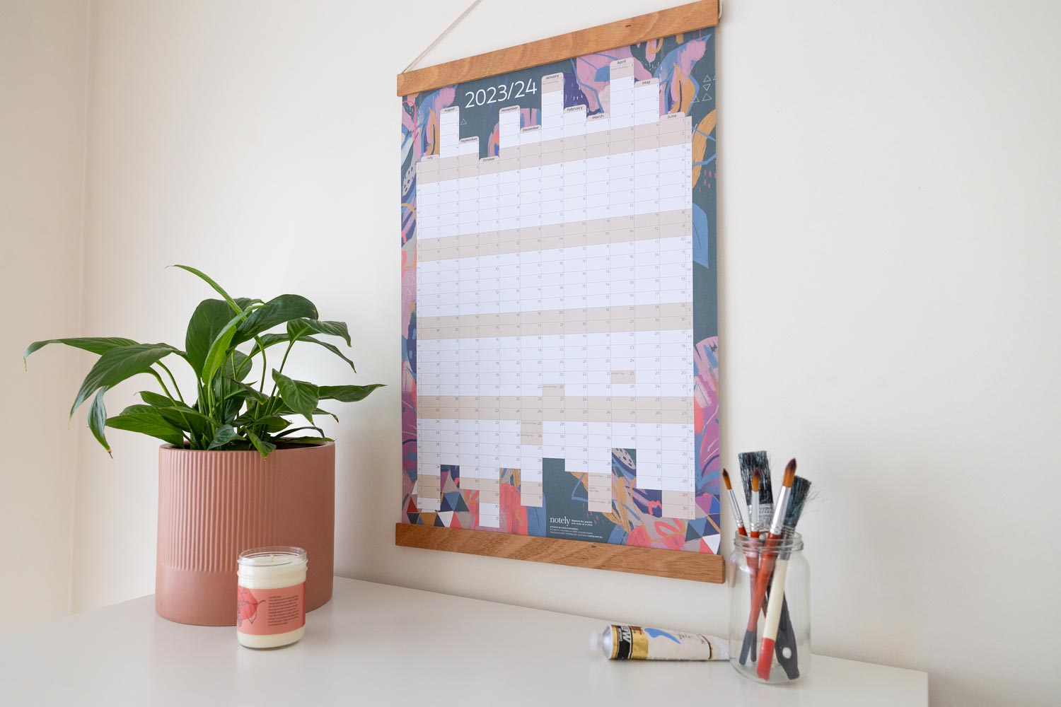 Notely Financial Year Wall Planner hanging on wall with pot plant, candle and paintbrushes in a jar