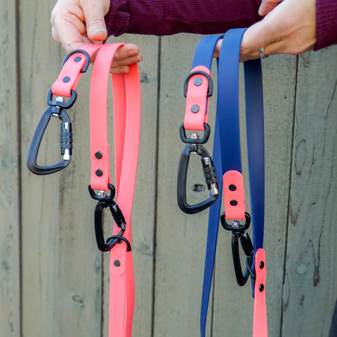 Two hands free biothane leashes with black carabiner hardware, being held up. the leash on the left is a coral pink color and the leash on the right is a midnight blue with pink coral accents.