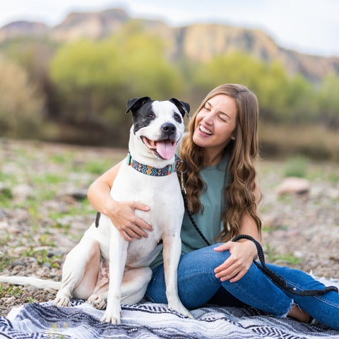 A woman with long brown hair sits on a southwestern style blanket in an open area with a desert landscape in the background.. A large white short haired dog with brown spots on its face sits next to her. The dog and person both have happy, relaxed expressions.