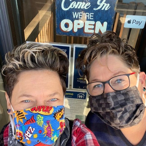 A close up photo of two white women with short brown hair and wearing cloth masks stand in front of a retail storefront, Maxwell's pet bar, with a sign in the window that says "come in, we're open."