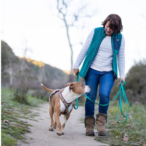 A woman with brown hair wearing jeans, a white shirt, and a teal fleece vest walks a large brown and white pit bull type dog along a trail in the desert at sunset. The dog is wearing a harness and a long teal leash and is looking up at the woman, and the woman is looking down at the dog, smiling.