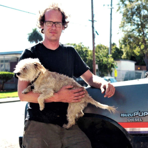 A man stands holding a white terrier dog next to an old Isuzu truck. 
