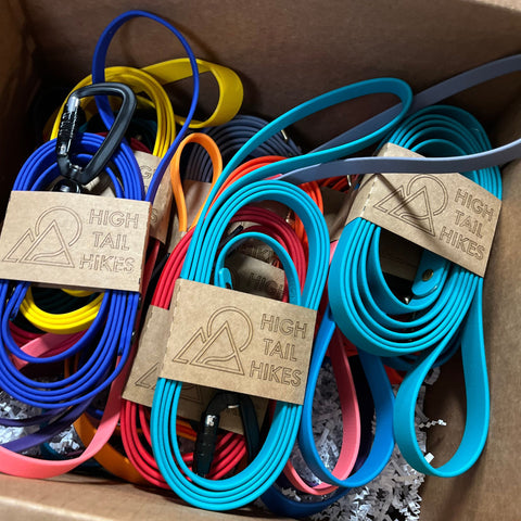 Several colorful waterproof BioThane leashes in shades of blue, yellow, red, and pink in karft card stock packaging sleeves that read "High Tail Hikes" stacked in a cardboard box.