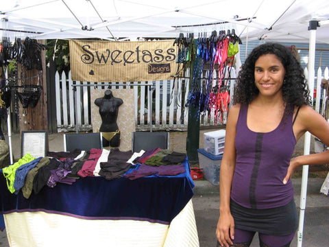 Nina J with her mid-2010s clothing line, Sweetass Designs.  She stands next to her vending table at a street festival, clothing and bikinis hang in the background