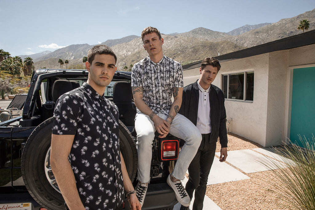 Image of three men posing in front of the back of a jeep, all of them wearing John Varvatos