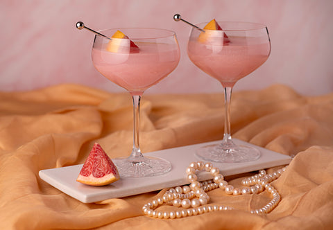 Two glass of grapefruit mocktails on a rectangle plate with a piece of fresh grapefruit and pink pearls displayed between the two glasses.