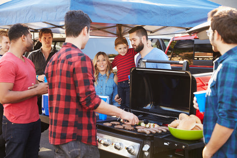 Group of people gathered outside around a man cooking on a grill.