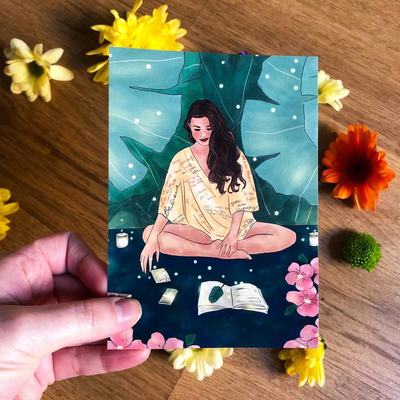 Noharanda illustrated postcard design of a woman sitting cross legged looking at affirmation cards and a journal, surrounded by plants and stardust