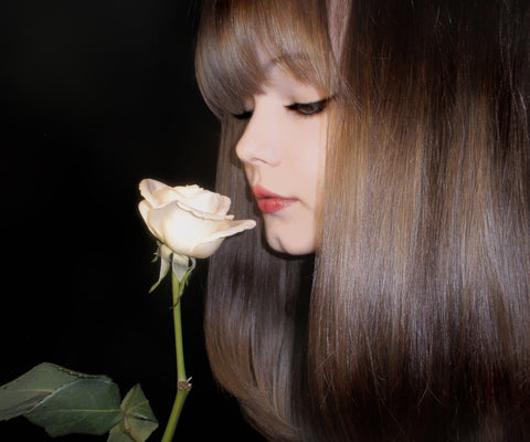 A beautiful young woman, with shiny brown hair, looking to her right and she is smelling a rose.