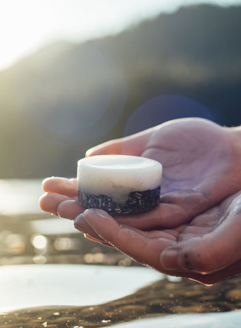 hands holding shampoo bar and conditioner bar above mountain lake water with sunset reflection.