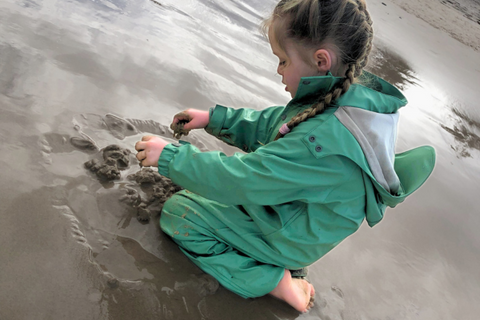 young girl in sage green puddle suit building the start of a sandcastle