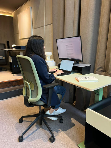 Persona sentada frente a un computador. Su escritorio es de altura ajustable y de color verde.