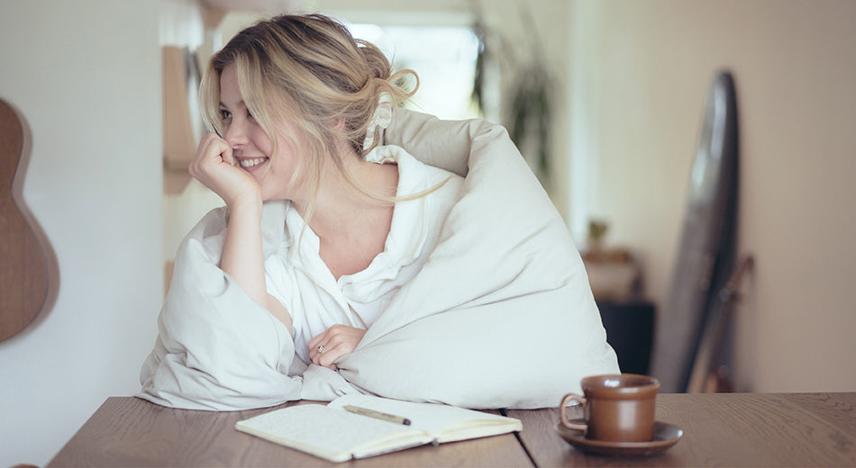 A woman sits at a table, wrapped in a duvet.  She is resting her chin in her hand and is smiling.  A book sits open on the table in front of her, next to a pen and a cup of coffee.  She is ready to start physically writing in her journal.