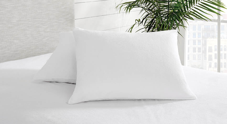 Two pillows with terry cloth pillow protectors sit on a bed dressed in a matching mattress protector.  A plant sits on the floor next to the bed.  There is a window in the background through which we can see an urban scene.