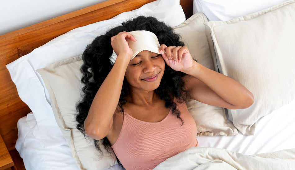 A young woman is lying in bed with two pillows behind her. She has a face mask on her forehead.