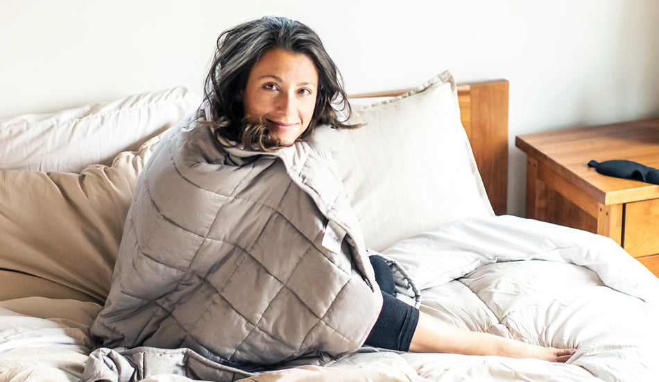 A woman is sitting on a bed with a weighted blanket wrapped around her.