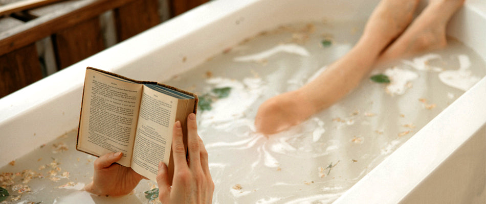 A person is taking a relaxing bath filled with aromatic herbs.  Only their hands and lower legs are shown.  They are reading a book.