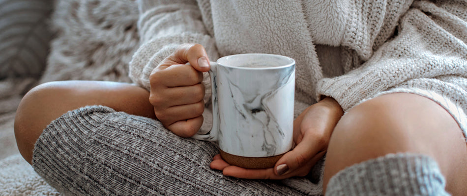 Close-up of a mug with warm milk, cradled in a woman’s hands.  She is wearing a cozy sweater and leg warmers.