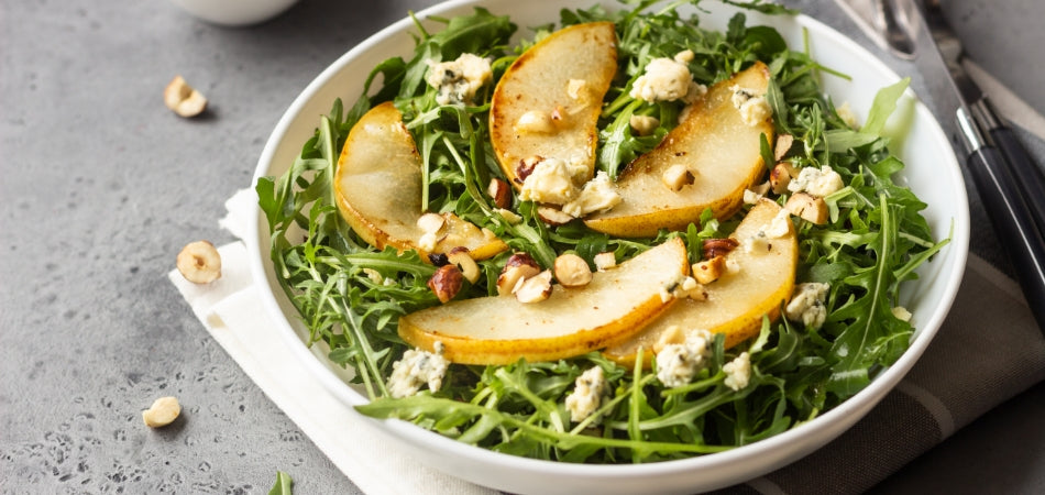 A white bowl with salad greens, sliced pears, nuts and cheese. The bowl is on a white napkin and a metal table.