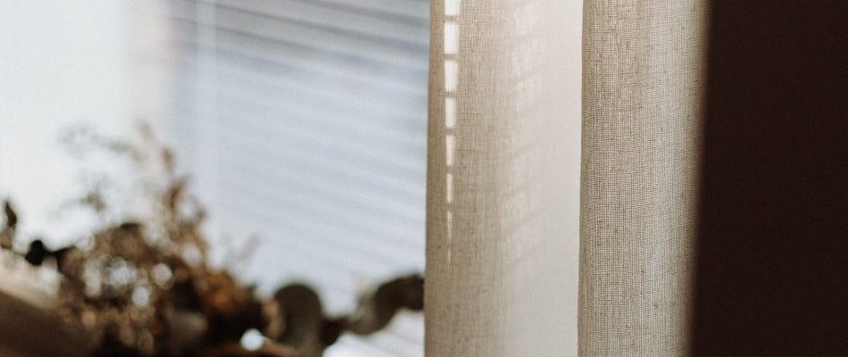 Beige curtain hanging next to a window which has blinds drawn.