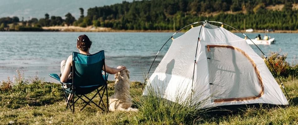 A woman sits on a camp chair at a campsite on a lake.  Her golden retriever dog sits next to her, and their tent is set up.  Both woman and dog are shown from behind, looking out over the water.