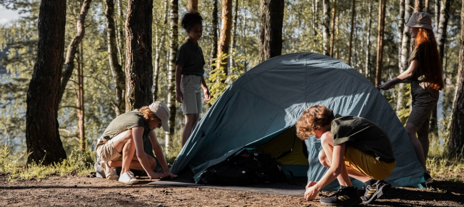 A group of young campers is setting up a tent at their campsite.  Dappled sunlight filters through the trees behind them.  We can see a lake in the background past the trees.