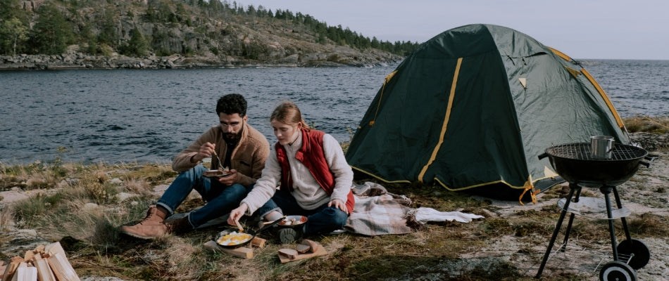Two campers sit on a blanket on the ground outside of their tent.  They are eating a meal, and their campsite is set up for cooking on a fire or grill. There is a lake in the background and the landscape is covered with rocks.