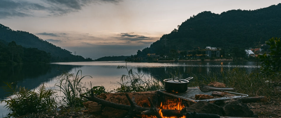 It is twilight on a lake.  A campfire slowly crackles and a pot of food sits steaming atop a grill.