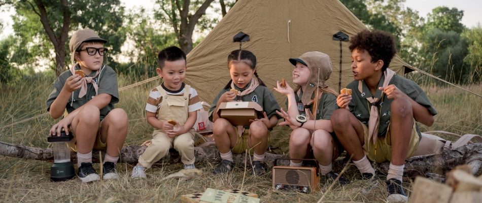 A group of young girl and boy scouts sit in camp outside of an old-fashioned tent.  They are talking, laughing, and eating snacks. Sunlight peeks through the trees behind them.