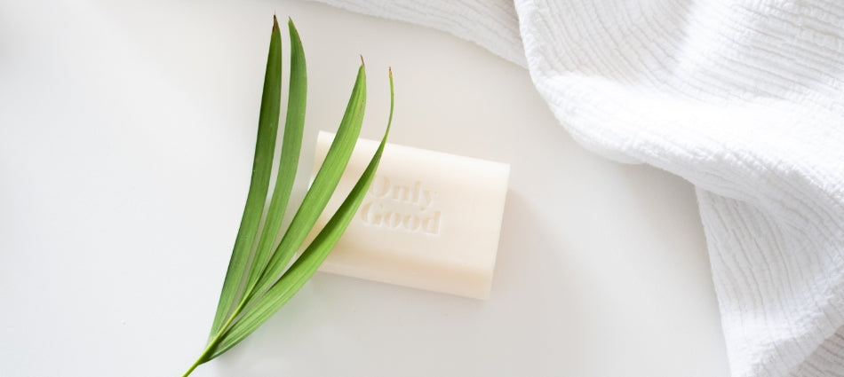 A bar of soap with the words Only Good, green leaves covering part of the soap and white fabric next to it.
