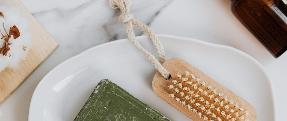 A white dish on a marble countertop. A nail brush with a hanging rope and a green bar of soap are on the white dish.