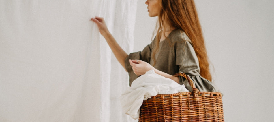 A woman is holding a wicker laundry basket on her left arm and her right hand is on a sheet which is hanging.