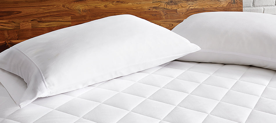 A close-up of two pillows on a bed.  They are dressed in white pillowcases.  The bed has a wood headboard and is dressed in only a white mattress pad.