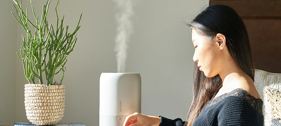 A woman sits on the side of a bed.  There is a nightstand next to her, and on it are a portable humidifier and a green potted plant.