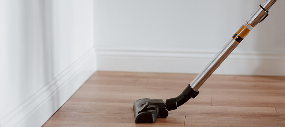 A compact, upright vacuum is shown in a corner, and it appears as though a person is using it to vacuum the wood floor. 