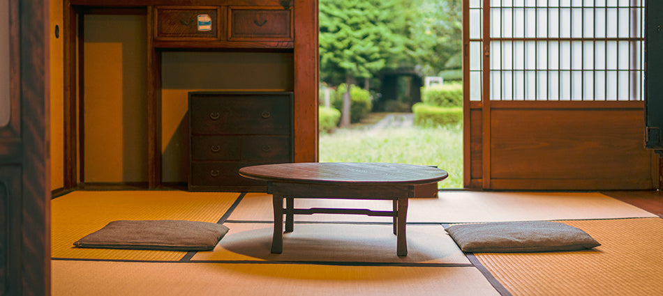A low table and two cushions sit in the middle of a traditional Japanese living room.  The floor is covered with tatami mats and the sliding door is open, showing a glimpse of a lush green yard.