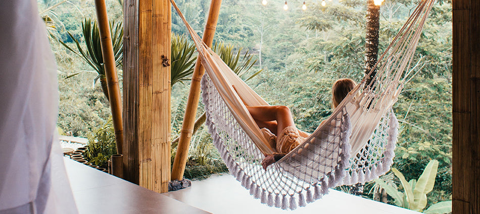 A woman reclines in a hammock, looking off into the distance.  The hammock is strung on a porch with bamboo and string light accents, and looks out over a jungle landscape.
