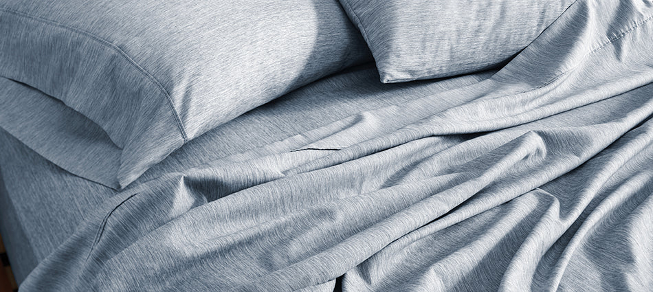 An unmade bed is shown in close-up.  The sheets are a heathered blue color and there are two pillows.