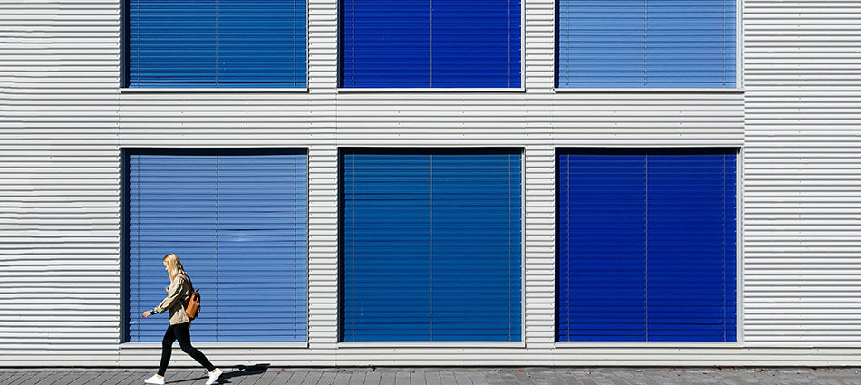 A pedestrian walks by a building façade that has six windows in it.  Each window has a different shade of blue horizontal blinds in it.