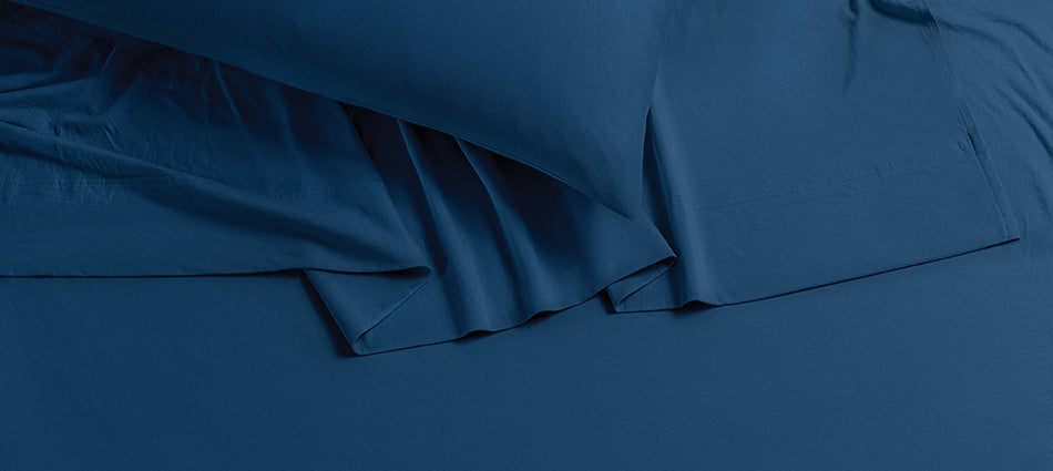 A close-up of a pillow with a rumpled bed sheet underneath.  The sheets are dark blue in color.