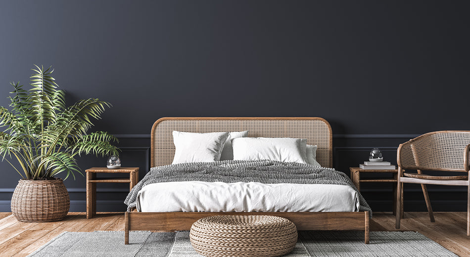 A bedroom scene is shown from the foot of the bed.  Furniture is all wicker or rattan, and colors are soft naturals.  The wall is a dark gray.